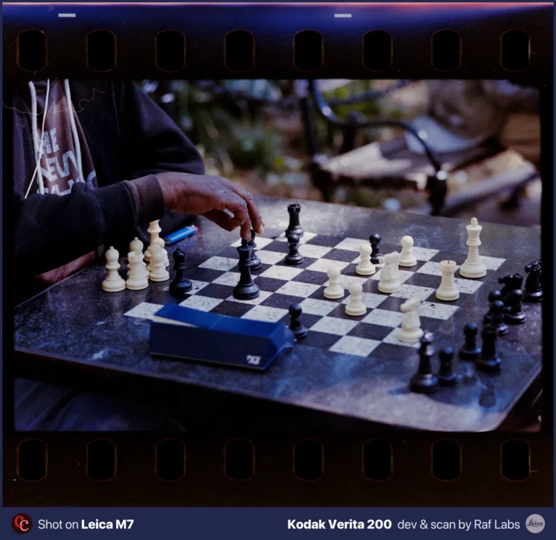 A person's hand reaches for a black chess piece on a marble-topped table during an outdoor game.