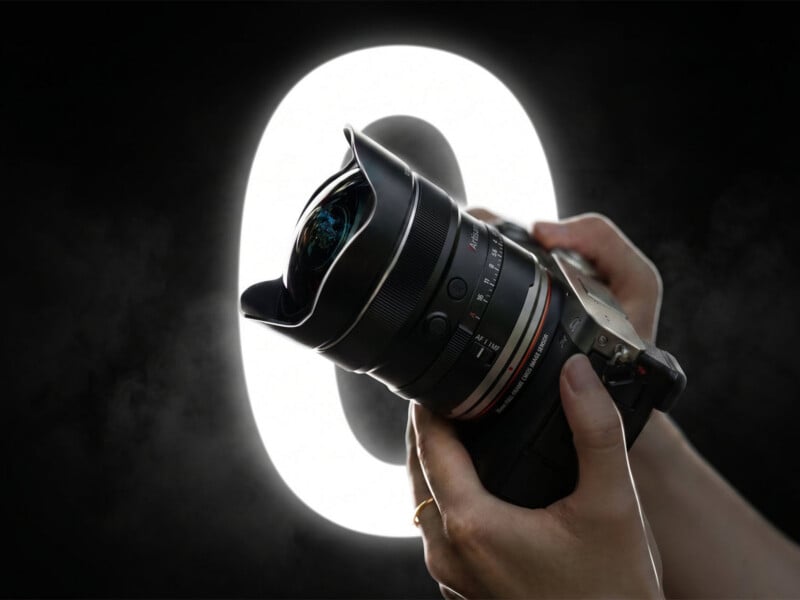 A person's hands hold a camera with a wide-angle lens in front of a glowing, circular light against a dark background.
