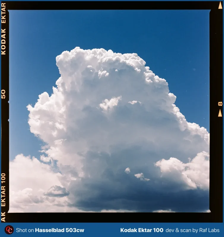 A large, white cumulus cloud with a dark, shadowed base stands against a clear blue sky, framed by film borders with text.