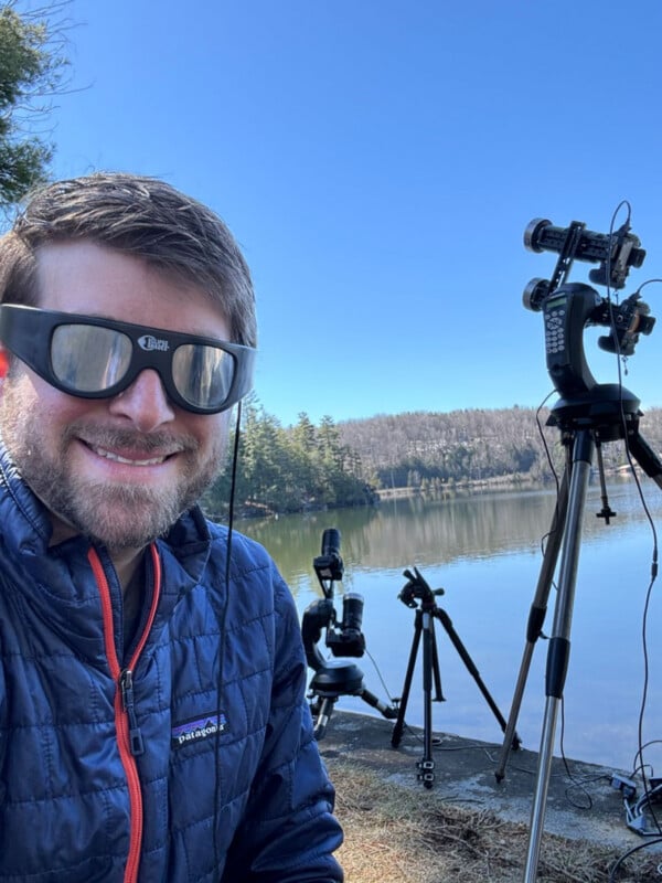A man wearing eclipse glasses smiles for a selfie outdoors by a lake, with two camera tripods set up behind him.