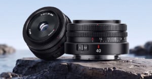 Two 7Artisans 40mm f/2.5 camera lenses rest on a dark, textured rock against a blurred blue background.