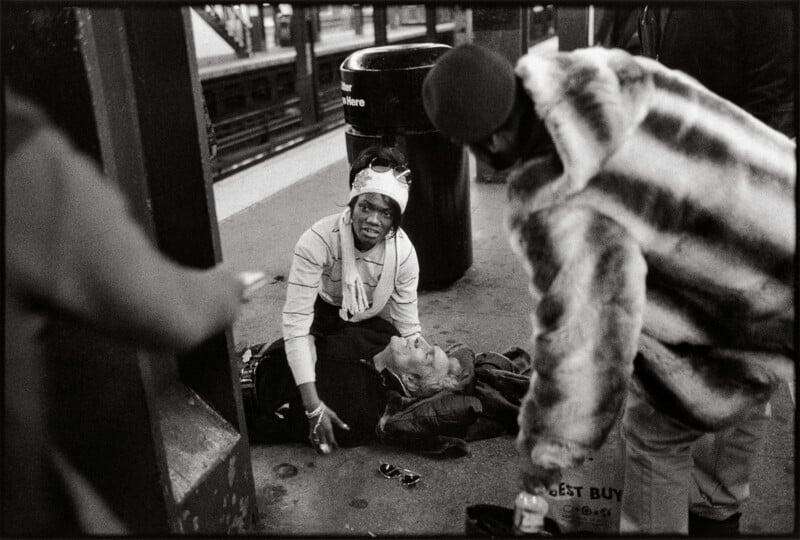 A woman kneels on a subway platform to assist a person lying on the ground, while a man in a fur coat stands nearby.
