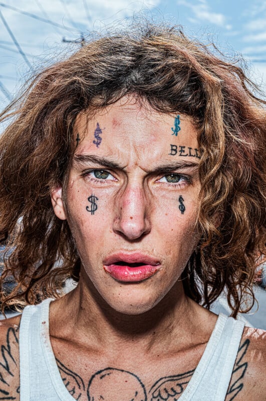 A close-up portrait of a person with curly brown hair, facial tattoos including dollar signs and the word "BELL," and a winged chest tattoo.
