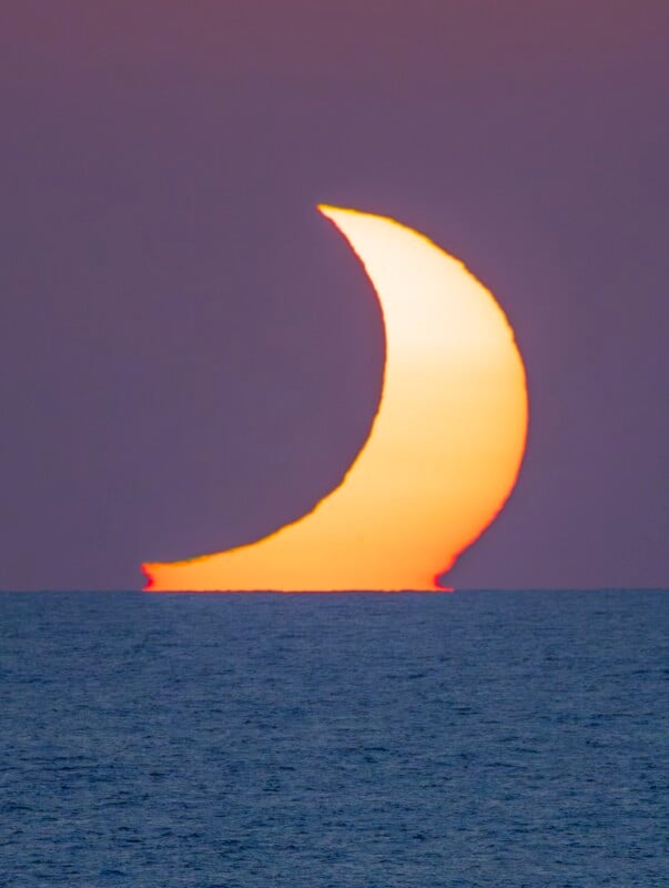 A crescent-shaped sun rises above a dark blue ocean horizon against a purple sky.