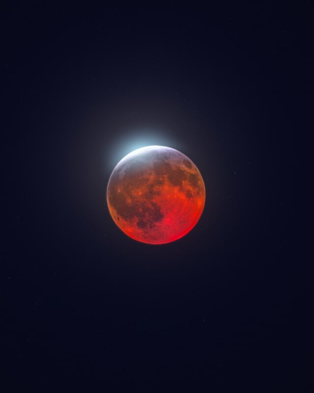 A lunar eclipse shows a reddish-orange moon against a dark, starry sky with a bright blue glow at the top edge.
