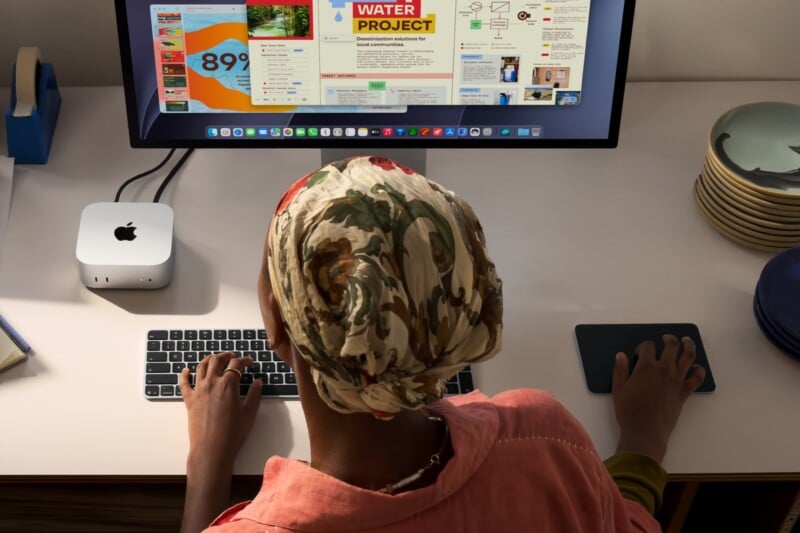 A person wearing a floral headscarf sits at a desk using a Mac Mini, a keyboard, and a trackpad to view a water project presentation on a large monitor.