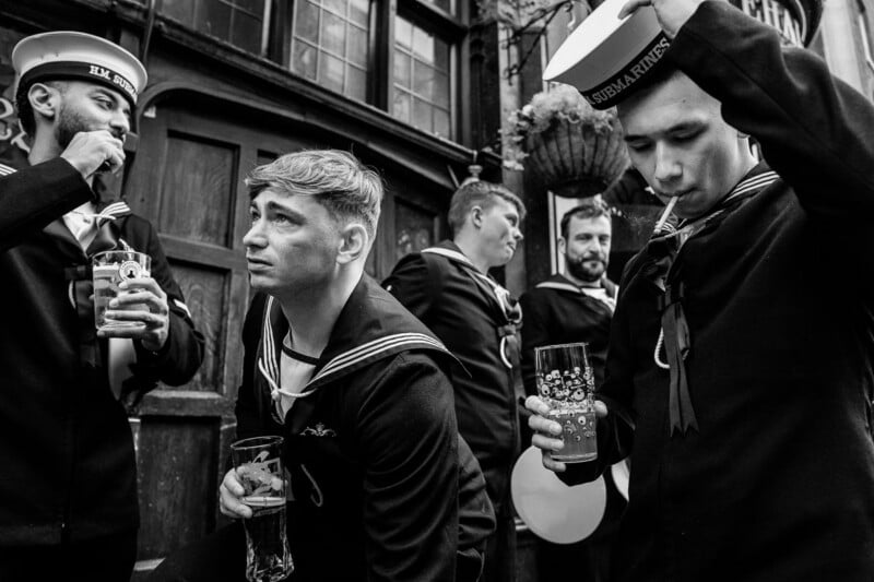 Five men in Royal Navy submarine service uniforms stand outside a building, some holding glasses of beer and one smoking a cigarette.