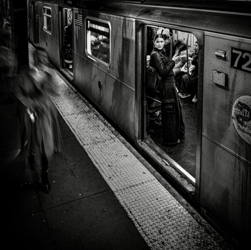 A black-and-white shot of a subway platform where a blurred figure walks past a train car with an open door, revealing a woman standing inside.