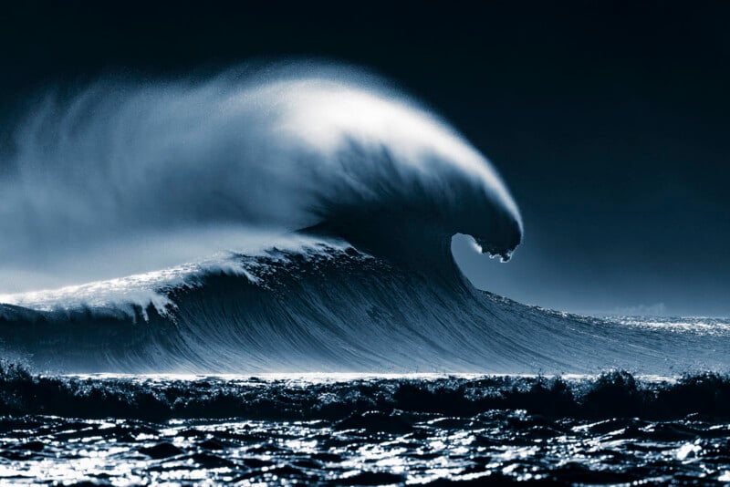 A large, dark blue ocean wave curls and breaks, with sea spray blowing off the top against a dark sky.