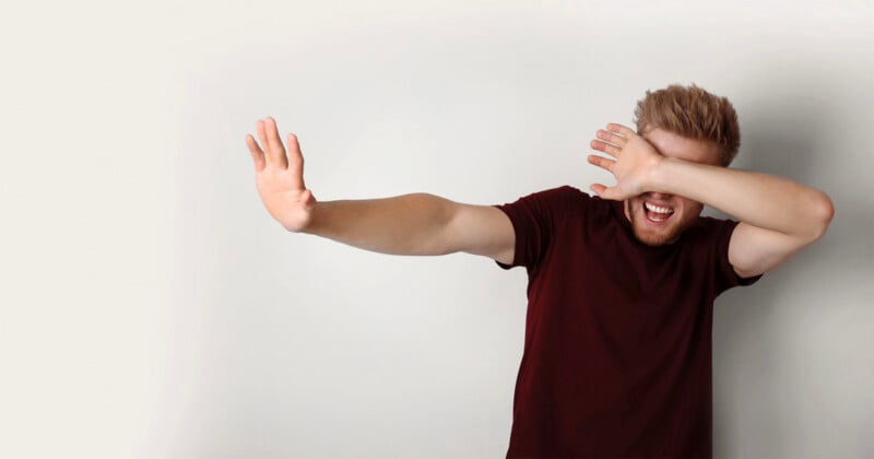 A man with reddish-brown hair and a beard wearing a maroon t-shirt holds one hand out in a stop gesture while covering his eyes with his other hand.
