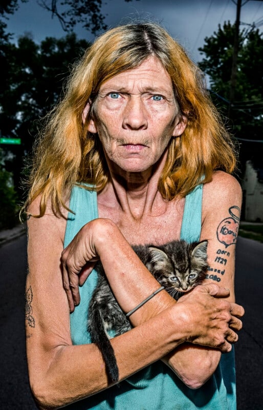 A woman with shoulder-length reddish-blonde hair and a teal tank top holds a small tabby kitten against her chest while standing outdoors.