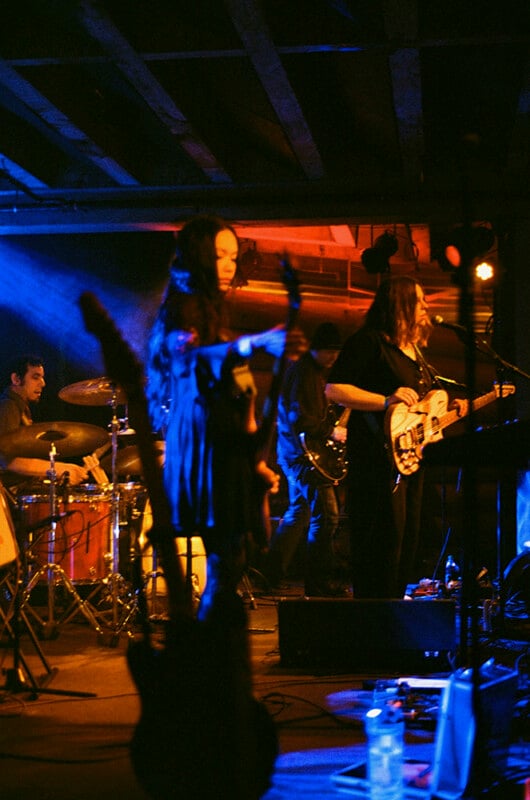 A band performs on a dimly lit stage with blue and orange lighting, featuring a drummer, a guitarist, and a bassist.