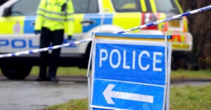 A blue and white police sign with a left-pointing arrow stands in the foreground, with a police car and an officer in a high-visibility jacket visible behind police tape in the background.