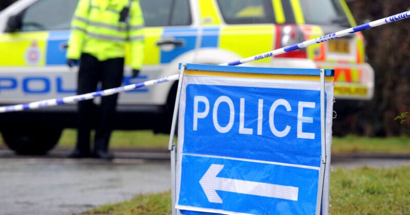 A blue and white police sign with a left-pointing arrow stands in the foreground, with a police car and an officer in a high-visibility jacket visible behind police tape in the background.