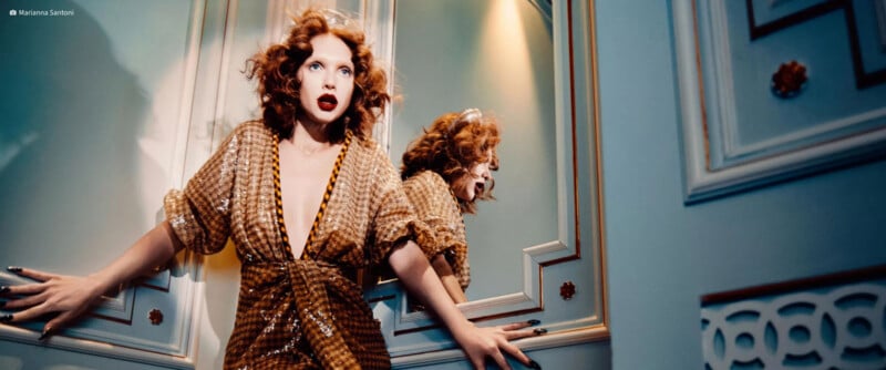 A woman with curly red hair and dark lipstick wears a sequined gold dress while posing in front of a mirror.