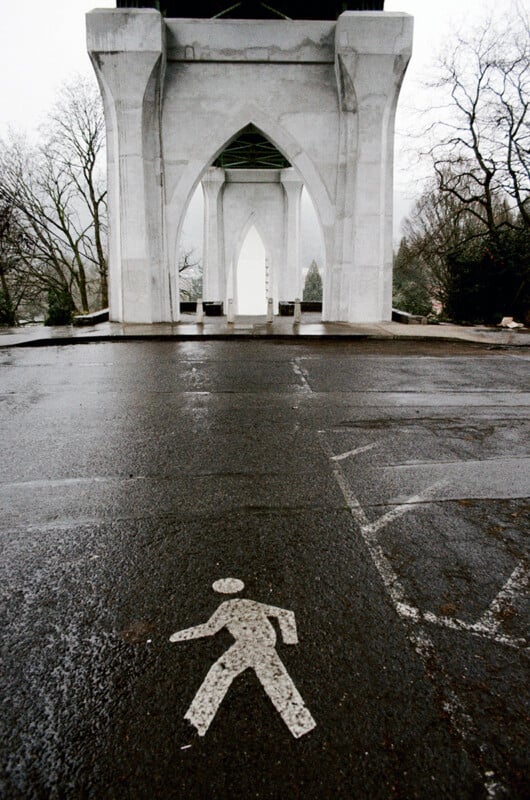 A pedestrian crossing symbol is painted on wet asphalt in front of large, arched concrete bridge supports.