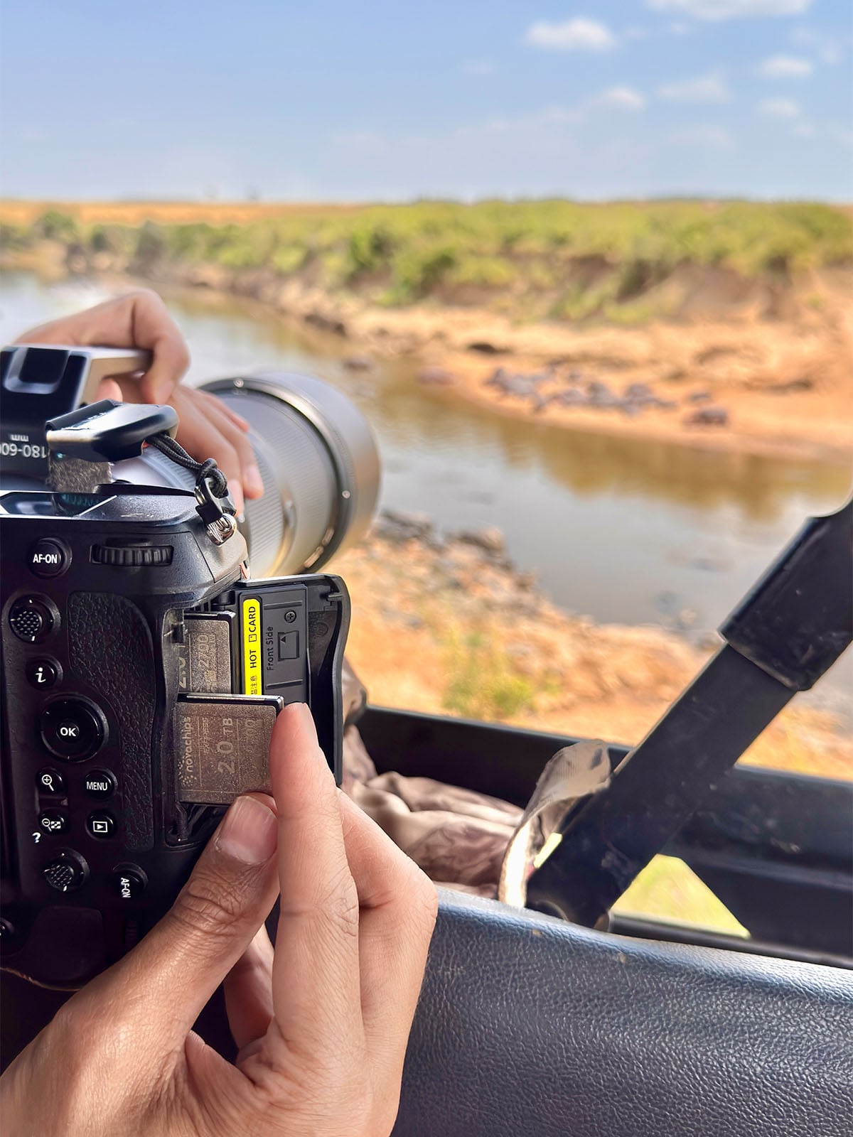 A person's hands insert a 2.0 TB memory card into the side slot of a professional camera while overlooking a riverbank with wildlife in the distance.