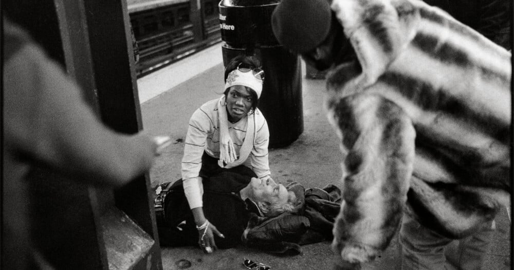 A woman in a white headband kneels on a subway platform, tending to a man lying on the ground as another person in a fur coat stands nearby.