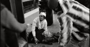 A woman in a white headband kneels on a subway platform, tending to a man lying on the ground as another person in a fur coat stands nearby.