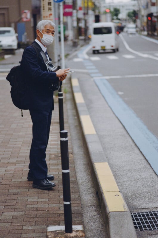 An older man in a suit and face mask stands on a sidewalk, holding a smartphone while looking toward the camera.