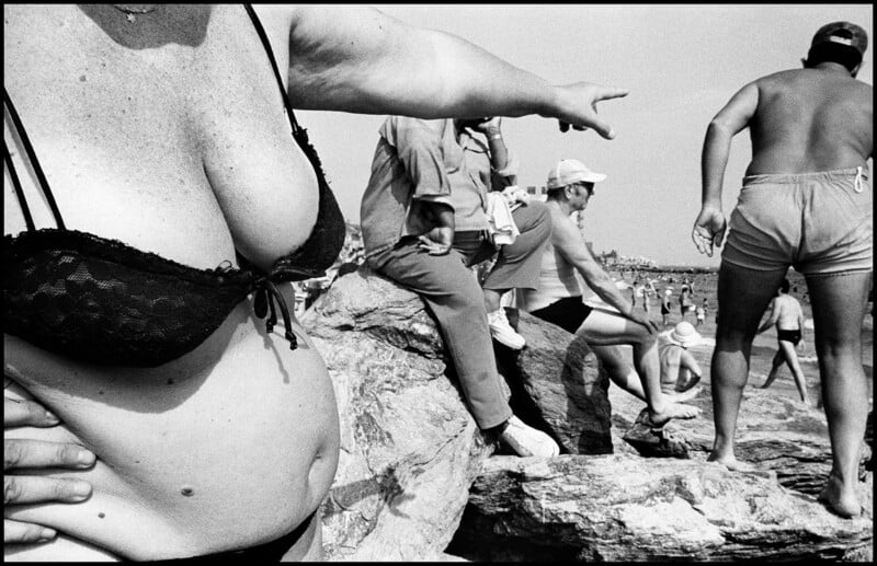 A close-up of a woman in a black lace bikini top points toward a beach scene where people sit on rocks and walk near the water.