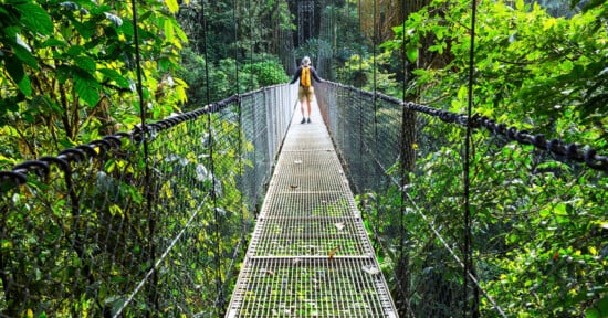 A person with a yellow backpack walks across a metal suspension bridge surrounded by a dense, green rainforest.