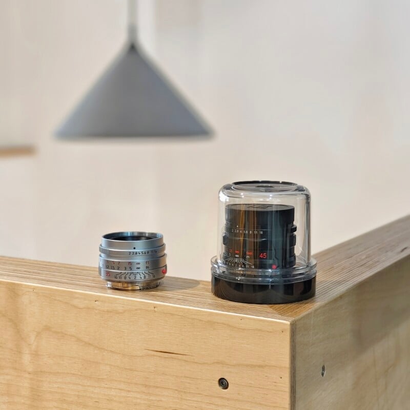 A silver camera lens sits next to a black camera lens encased in a clear plastic dome on a wooden surface.