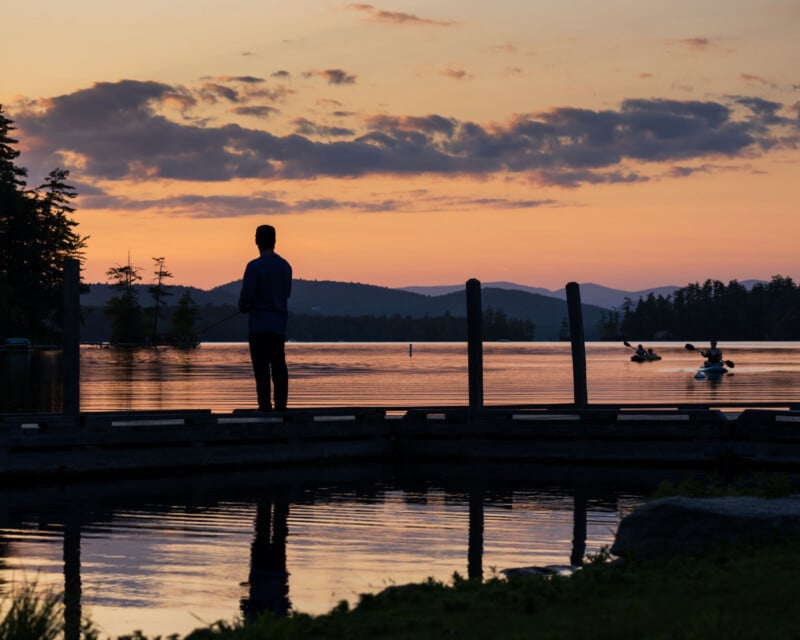 A person stands on a wooden dock fishing at sunset, while two people paddle kayaks in the distance on a calm lake.