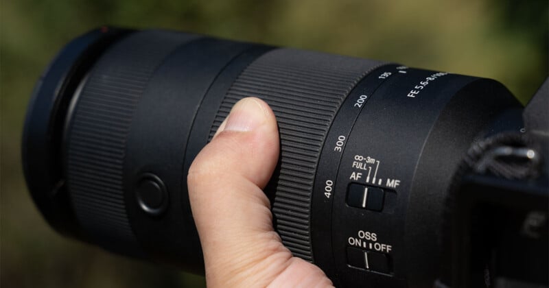 A close-up view of a hand holding a black telephoto camera lens, showing zoom markings from 135 to 400, focus limit switches, and an optical stabilization switch.