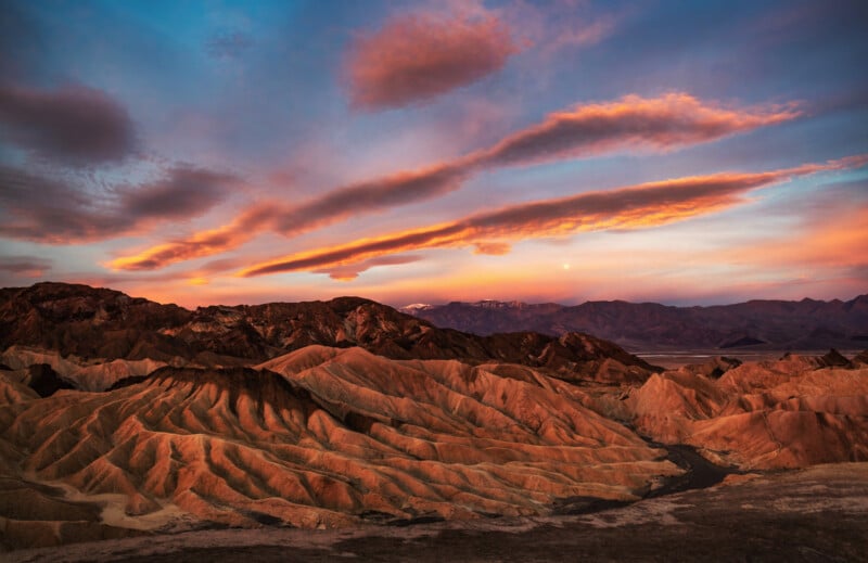 A dramatic sunset illuminates the ridged, golden-brown badlands of a desert landscape beneath a sky filled with colorful clouds.