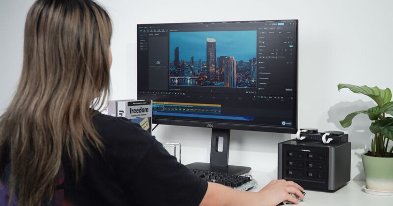 A person with long brown hair sits at a desk editing a video of a city skyline on a computer monitor, with a multi-bay storage device and a potted plant nearby.
