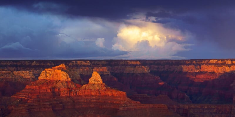 A dramatic sunset illuminates the layered rock formations of a deep canyon under a dark, stormy sky with bright clouds.