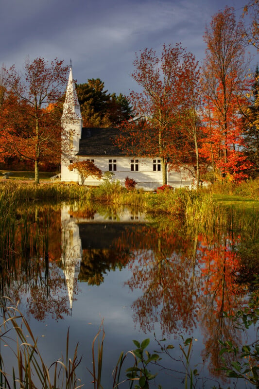 A white church with a steeple stands beside a pond reflecting the building and surrounding autumn trees with vibrant orange and red foliage.
