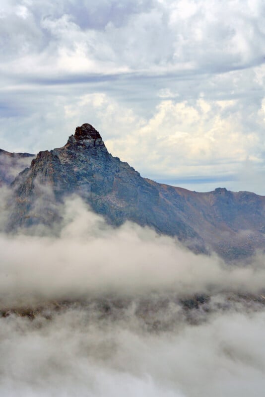 A jagged, rocky mountain peak rises above a thick layer of low-hanging clouds under a cloudy sky.