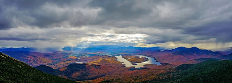 A panoramic view of a mountain valley during autumn, featuring a lake surrounded by colorful foliage under a dramatic, cloudy sky.