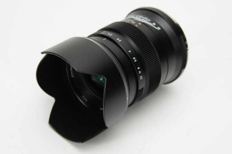 A black camera lens with a petal-shaped lens hood, showing aperture markings from 2.4 to 22, resting on a white surface.