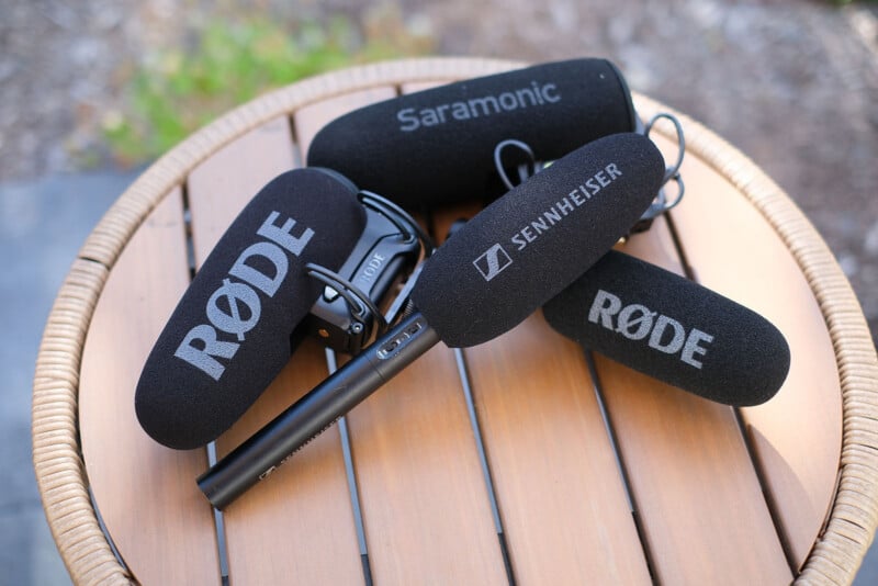 Three microphones with foam windscreens labeled RØDE, Saramonic, and Sennheiser rest on a round, slatted wooden table.