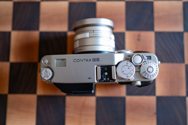 A top-down view of a silver Contax G2 camera with a lens attached, resting on a wooden checkerboard surface.