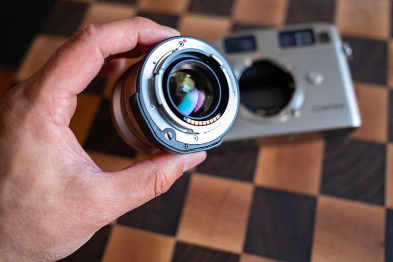 A hand holds a camera lens with visible electronic contacts, positioned in front of a silver camera body resting on a checkered wooden surface.