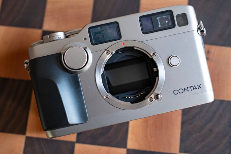 A silver Contax camera body with a lens mount, viewfinder windows, and a black grip, resting on a wooden checkered surface.