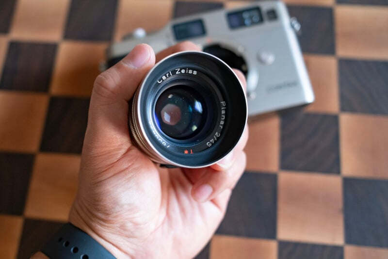 A hand holds a Carl Zeiss Planar 2/45 lens in front of a silver Contax camera body resting on a checkered surface.