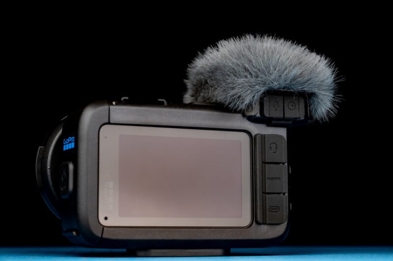 A black action camera with a fuzzy gray windscreen attached to the top, set against a black background on a blue surface.