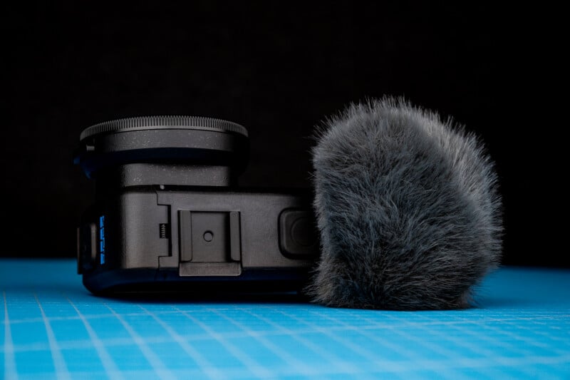 A black action camera with a circular lens filter sits next to a fluffy grey wind muff on a blue grid surface against a black background.