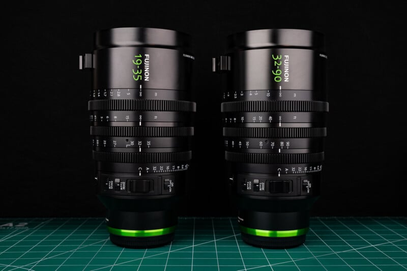 Two Fujinon cine zoom lenses, a 19-35mm and a 32-90mm, stand upright on a green cutting mat against a black background.