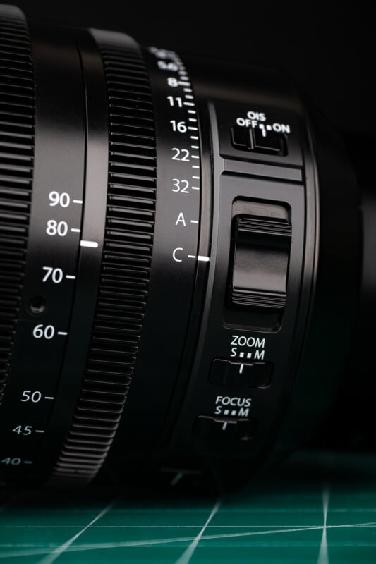 A close-up of a camera lens barrel featuring aperture settings, an OIS switch, a zoom toggle, and focus mode switches.