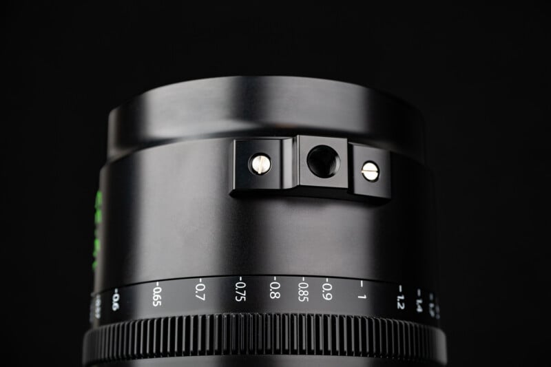 A close-up of a black camera lens barrel featuring a focus ring with distance markings and a mounting point with a threaded hole secured by two screws.