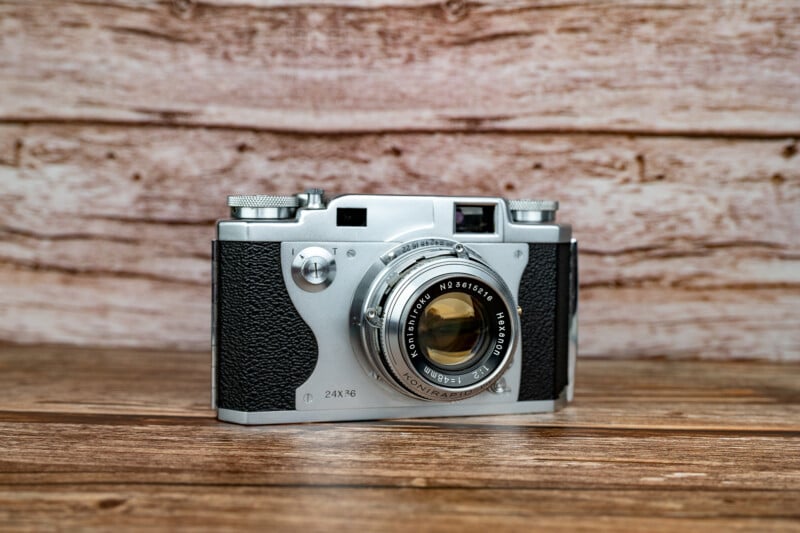 A vintage Konishiroku Hexanon 35mm rangefinder camera sits on a wooden surface against a wood-paneled background.
