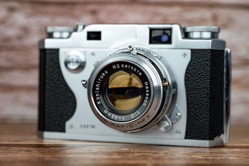 A vintage silver and black Konishiroku Hexanon 48mm f/2 film camera sits on a wooden surface against a blurred brick background.
