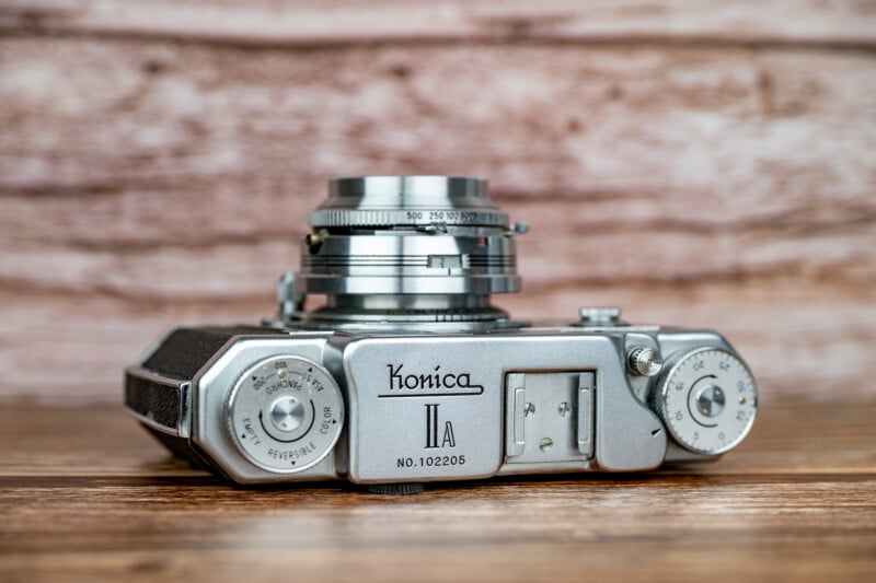 A silver Konica IIA rangefinder camera sits on a wooden surface against a light-colored, textured background.
