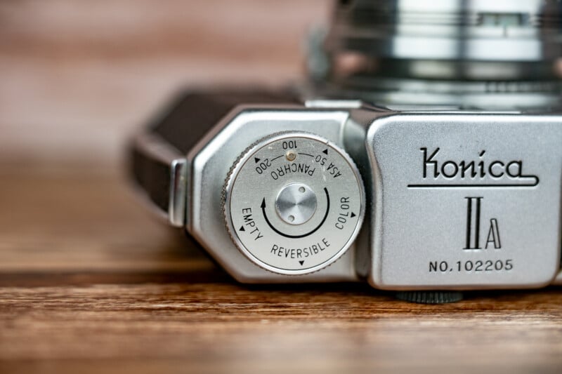 A close-up of the top plate of a silver Konica IIA camera, showing a film reminder dial labeled with ASA settings and film types, resting on a wooden surface.